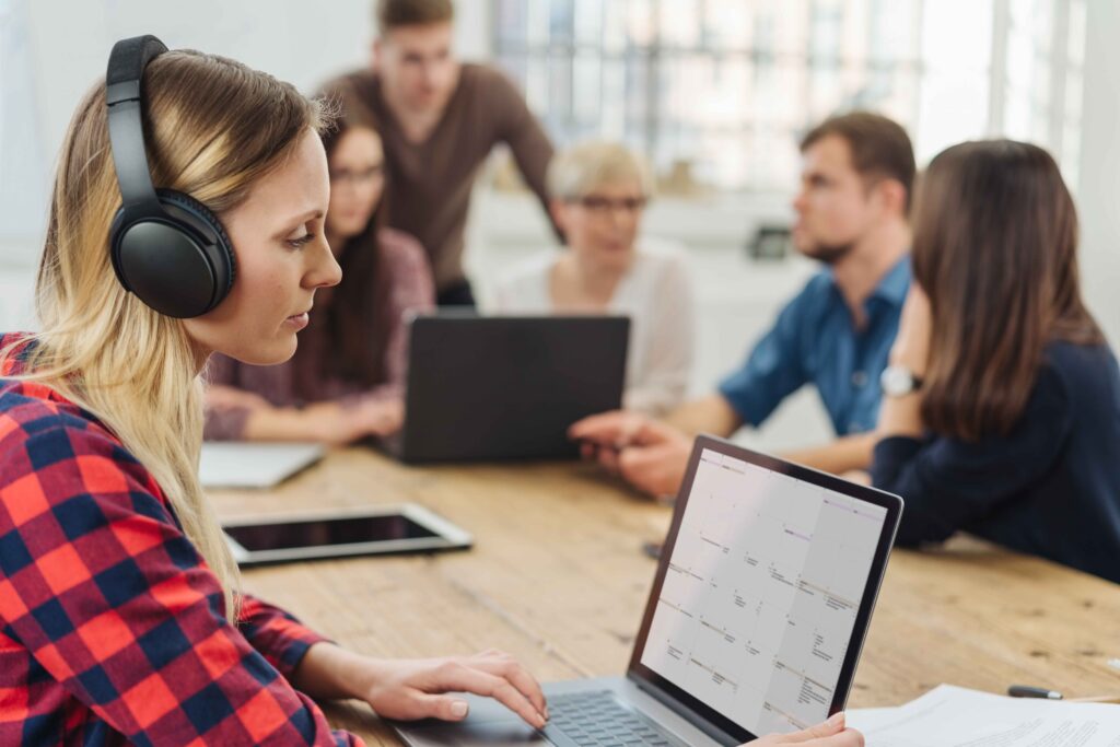 Vrouw werkt op kantoor achter een laptop met koptelefoon op. Er zitten pratende collega's om haar heen.